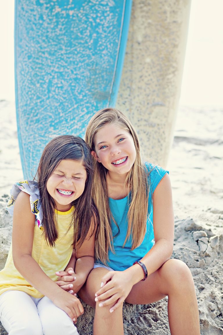 Two girls with metal braces laughing together on the sand in front of two surfboards, Hulse Orthodontics, Carlsbad CA