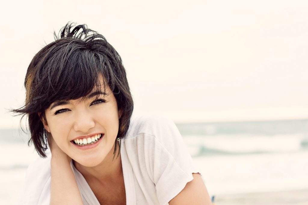Smiling young woman with short dark hair leaning forward on a beach, Hulse Orthodontics, Carlsbad CA