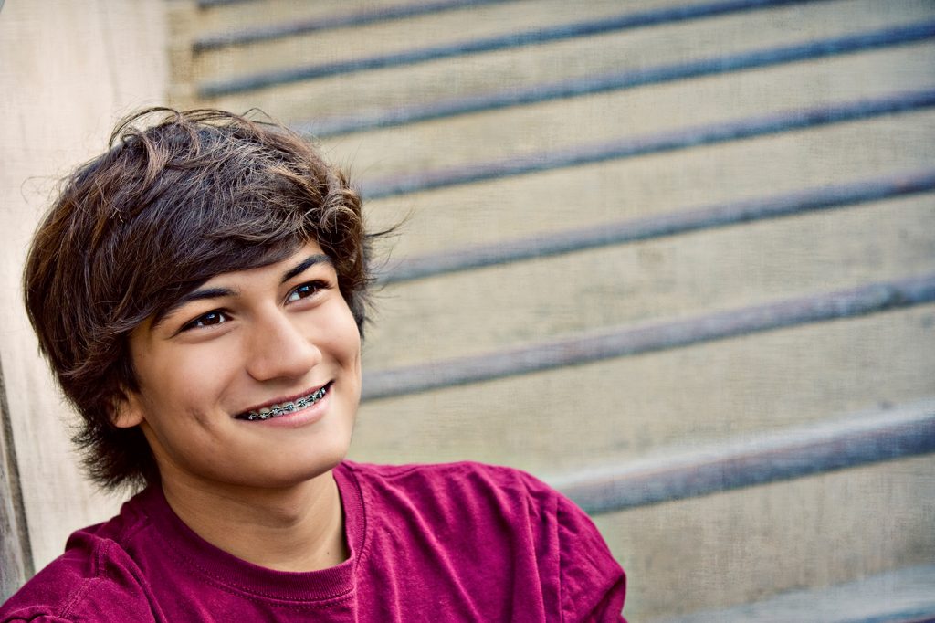 Smiling teenage boy with metal braces in a magenta T-shirt, sitting beside outdoor concrete steps, Hulse Orthodontics, Carlsbad CA