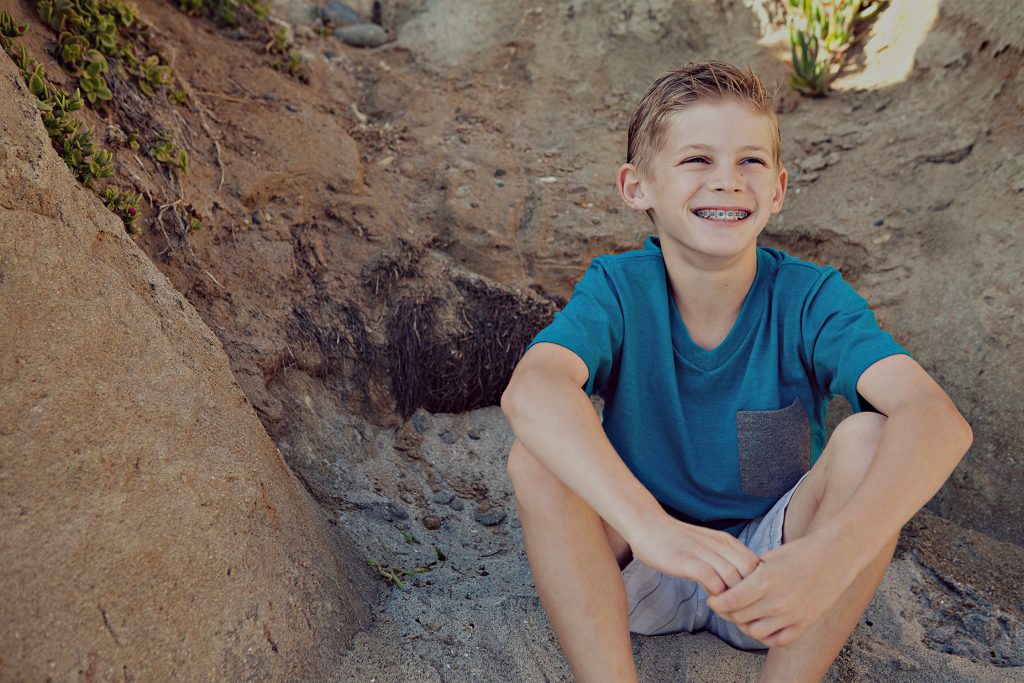 Smiling boy with metal braces in a teal T-shirt, sitting in the sand below a sandy bluff, Hulse Orthodontics, Carlsbad CA