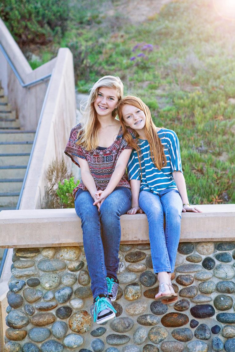 Two smiling girls with metal braces sitting side by side on a stone wall beside an outdoor stairway, Hulse Orthodontics, Carlsbad CA