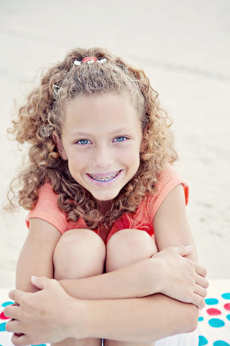Smiling girl with curly hair and metal braces with orange bands, hugging her knees on a beach, Hulse Orthodontics, Carlsbad CA