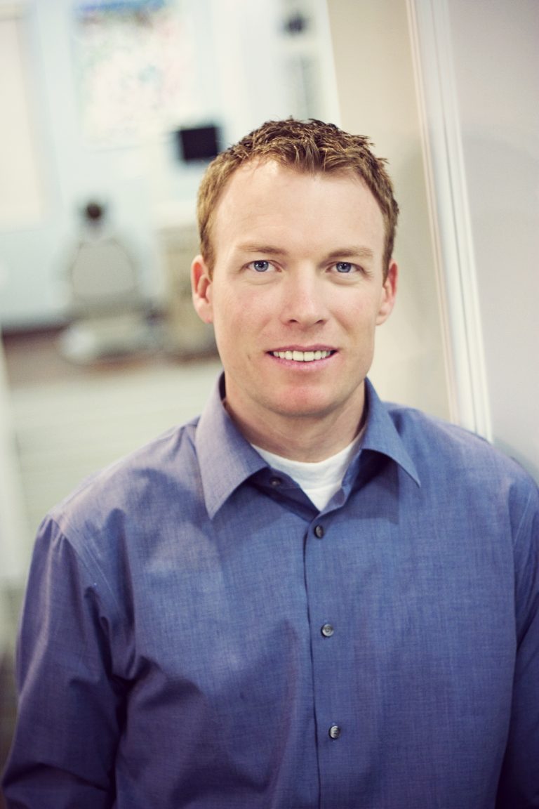 Dr. Cameron Hulse, specialist orthodontist, smiling in a blue button-down shirt with a treatment chair blurred behind him, Hulse Orthodontics, Carlsbad CA