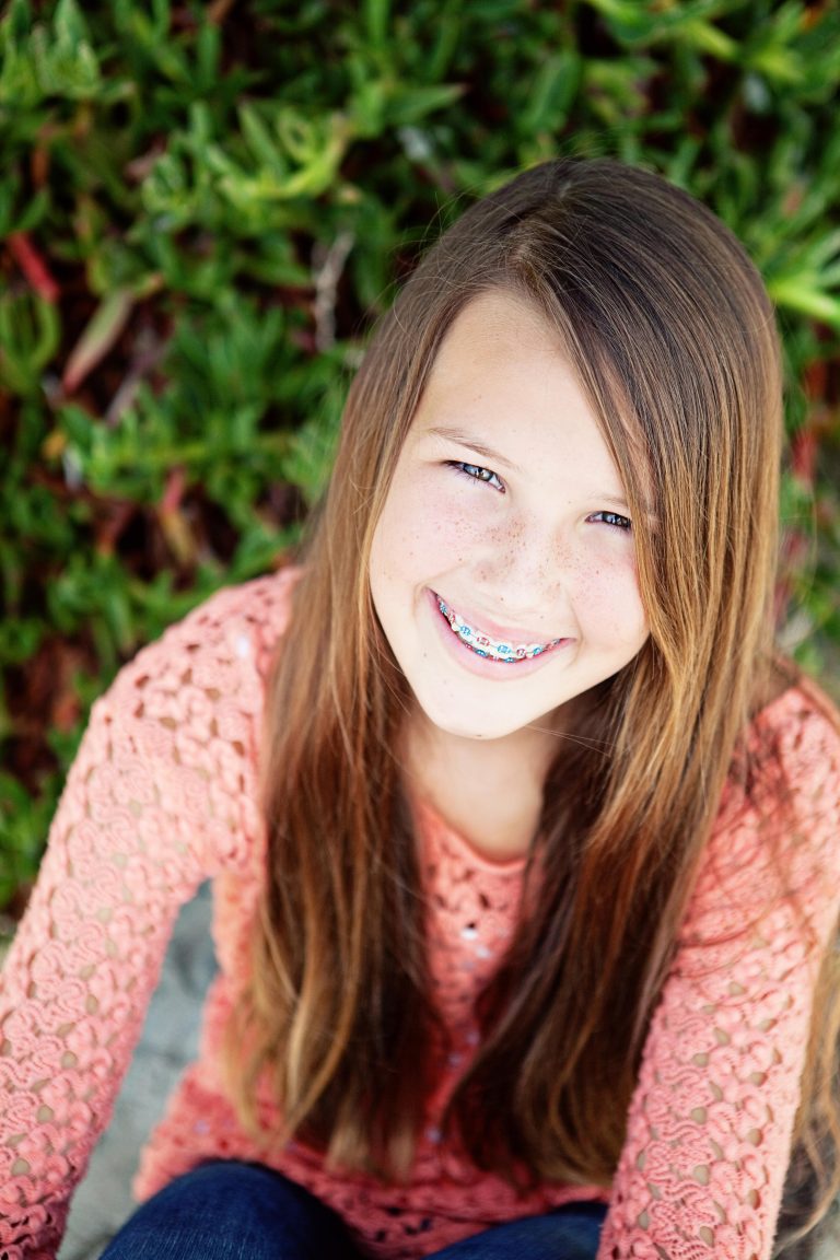 Smiling girl with metal braces with pink and blue bands, in a coral lace top in front of green plants, Hulse Orthodontics, Carlsbad CA