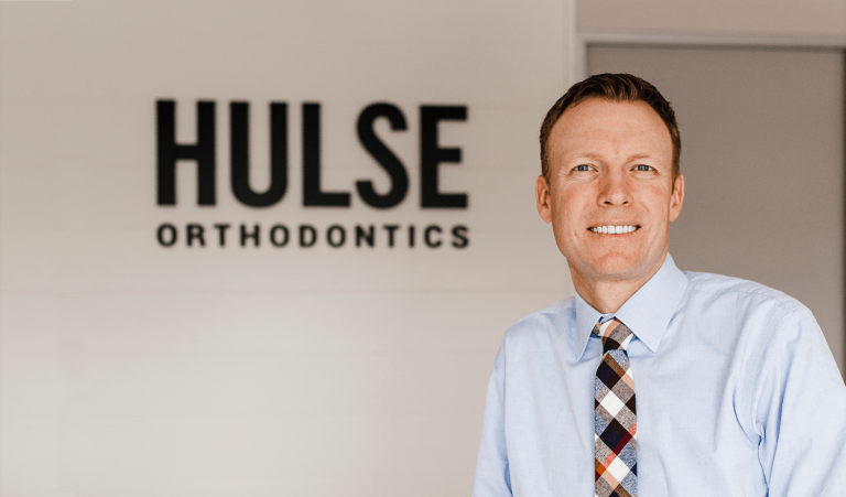 Dr. Cameron Hulse smiling beside the Hulse Orthodontics sign in the office, Hulse Orthodontics, Carlsbad CA