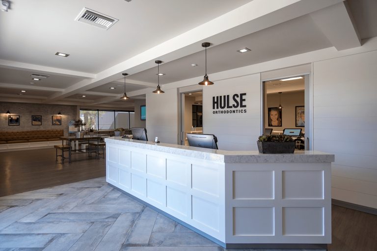 White reception desk under a Hulse Orthodontics wall sign, with pendant lights and a seating area to the left, Hulse Orthodontics, Carlsbad CA
